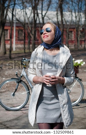 A young woman with a cup of coffee resting with a bicycle in early spring in a park, country style
