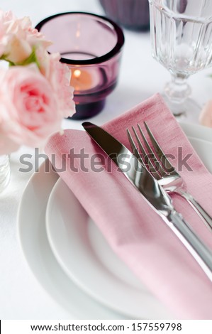 Beautiful festive table setting with roses, candles, shiny new cutlery and napkins on a white tablecloth.
