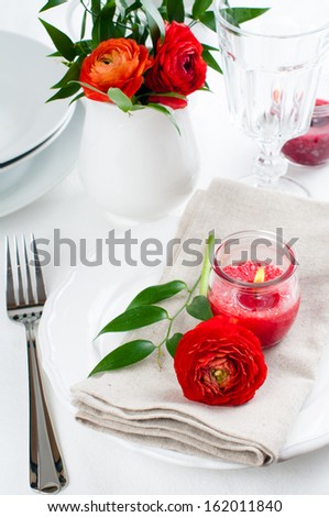 Festive dining table setting with red buttercup flowers, candles, napkins and shiny new cutlery in white.