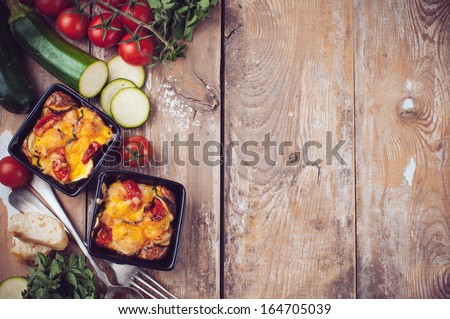 Two dishes of vegetable casserole with cheese, zucchini, cherry tomatoes and oregano on a rustic wooden board with vintage cutlery, food background, home cooking
