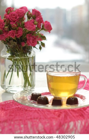 Glass cup with green tea, chocolates and pink roses on the table by the window