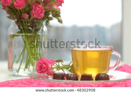 Glass cup with green tea, chocolates and pink roses on the table by the window