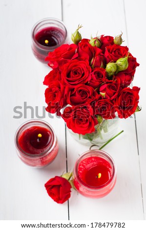 Small bouquet of red roses in a vase and candles on a white wooden table, rustic style