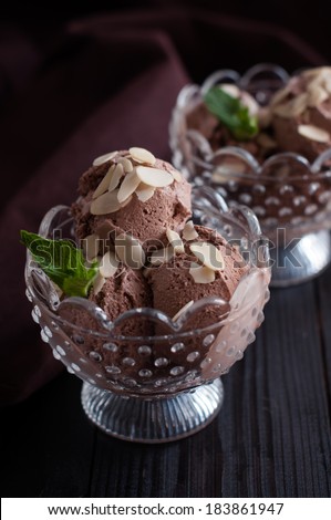 Two portions of delicious chocolate ice cream with almonds and mint leaf in crystal bowls and dark brown cloth on a wooden board, appetizing dessert, food closeup.