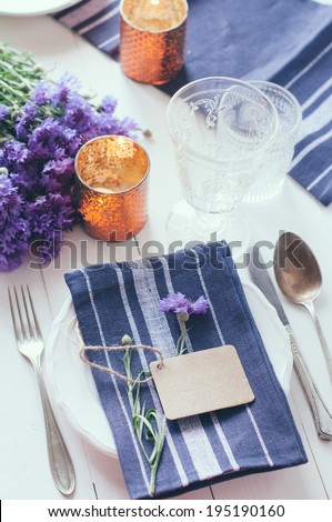 Vintage home table setting with blue napkins, antique cutlery and purple cornflowers on white wooden table. Blank cardboard tag and an old key