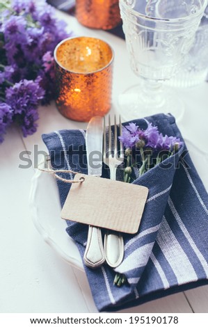 Vintage home table setting with blue napkins, antique cutlery and purple cornflowers on white wooden table. Blank cardboard tag.