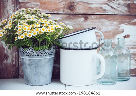Flowers in a tin can, glass bottles and vintage enamel mugs on wooden background, cozy home rustic decor, cottage living