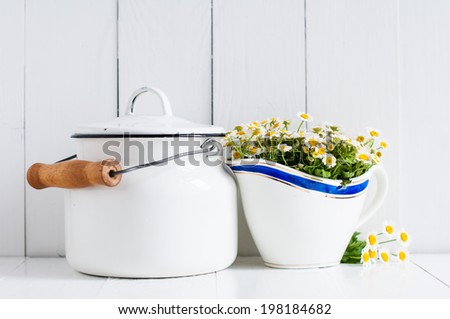 Chamomile flowers in vintage ceramic gravy boat and enamel milk can on white wooden background, home rustic decor