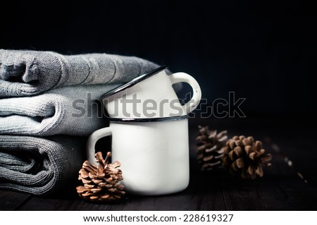 A stack of gray knitted winter sweaters, enamel mugs and fir cones on a dark background, rustic decor