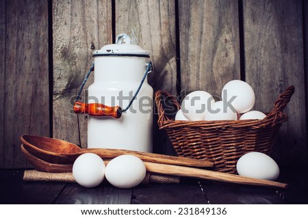 Country kitchen Still Life, enamel milk can, eggs in a wicker basket and wooden spoons on a background of the old board, grunge style