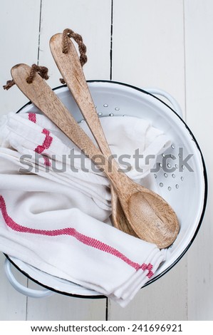 Rustic kitchen decor, enameled colander, wooden spoons and linen towels, dishes on a white board