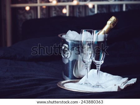 A bottle of chilled champagne in an ice bucket and two glasses on a bed, dark tones