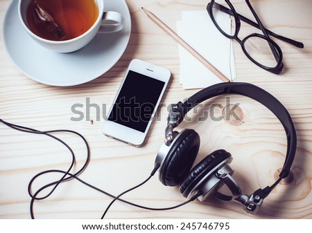 Paper records, cup of tea, mobile phone, headphones, glasses and pencil on wooden office desktop, hipster style