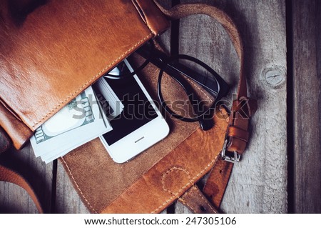 Optical glasses, money and smartphone in an open leather hipster\'s bag on a wooden board background.