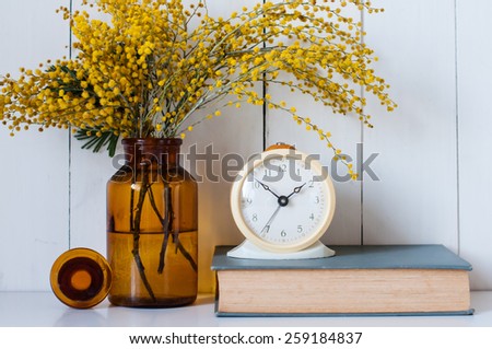 Home decor, mimosa yellow spring flowers in a vintage bottle, book and alarm clock on the white wall background