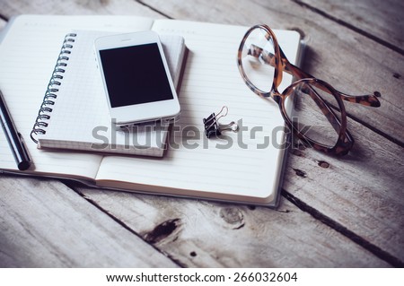 Hipster home office tabletop: papers and notebooks, reading glasses, smart phone, pen on an old wooden board background. Vintage lifestyle.
