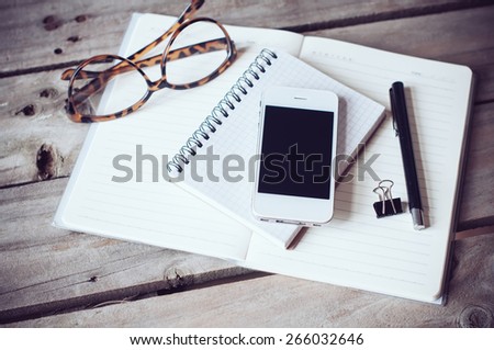 Hipster home office tabletop: papers and notebooks, reading glasses, smart phone, pen on an old wooden board background. Vintage lifestyle.