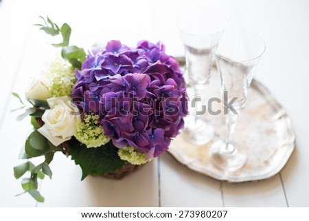 Big bouquet of fresh flowers, purple hydrangeas and white roses in a wicker basket, wine glasses and rustic wedding decor on a table, vintage style