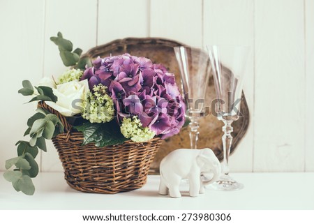 Big bouquet of fresh flowers, purple hydrangeas and white roses in a wicker basket, wine glasses and rustic wedding decor on a shelf in the interior, vintage style