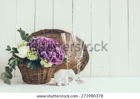 Big bouquet of fresh flowers, purple hydrangeas and white roses in a wicker basket, wine glasses and rustic wedding decor on a shelf in the interior, vintage style