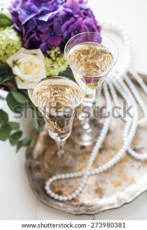 Big bouquet of fresh flowers, purple hydrangeas and white roses in a wicker basket, two glasses of champagne and vintage wedding decor with pearls on a table