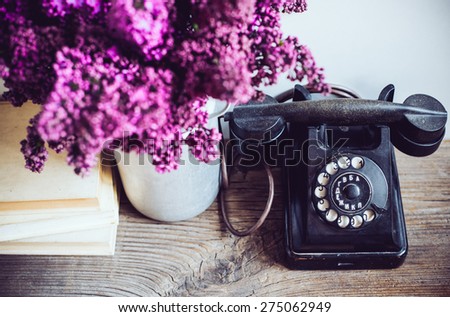 Home interior decor, bouquet of lilacs in a vase, a vintage rotary phone and books on rustic wooden table, on a white wall background