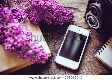 Books, notebooks, vintage camera, white smart phone and lilac flowers on an old wooden board background, hipster lifestyle arrangement