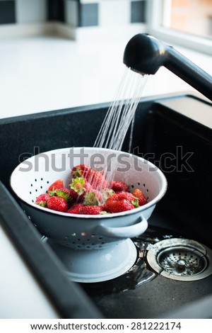 Fresh ripe organic strawberries in a white enameled colander under running water in the kitchen sink.