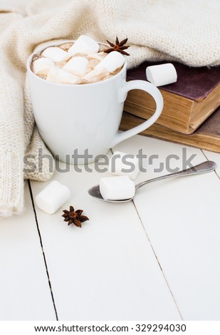 Cozy winter home background, cup of hot cocoa with marshmallow, old vintage books and warm knitted sweater on white painted wooden board background.
