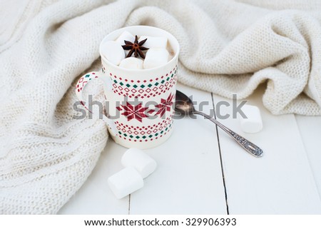 Cozy winter home background, cup of hot cocoa with marshmallow and star anise, old vintage books and warm knitted sweater on white painted wooden board background.