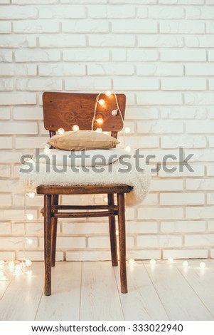 A stack of white and beige pillows and blankets with string lights on vintage wooden chair. Cozy interior details, soft and warm home decor.