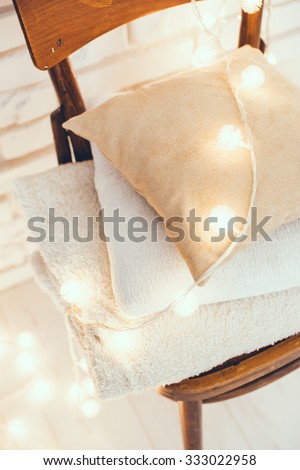A stack of white and beige pillows and blankets with string lights on vintage wooden chair. Cozy interior details, soft and warm home decor.