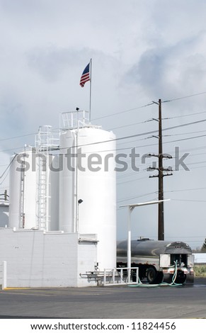 Vertical image of a tanker filling his truck from tall white silos.  An American flag is flying from the top of the near silo.