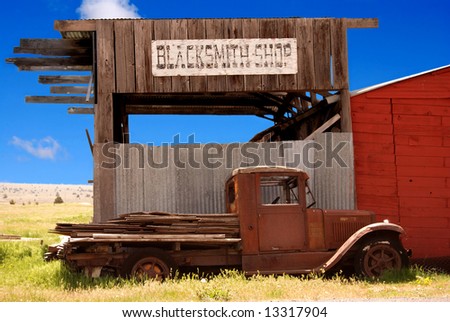 Horizontal image of an old rusted truck in front of a decaying blacksmith shop.  Old west image against a bright blue sky.