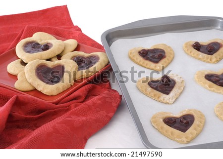 Unbaked sheet of cherry heart cookies next to a red plate of baked cookies setting against a red napkin.