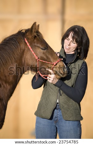 Pretty dark haired woman standing with her sorrel (chestnut) quarter horse foal against an out of focus barn.
