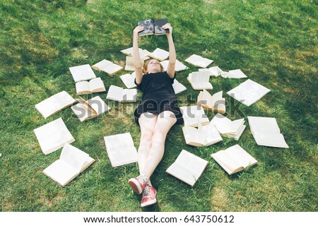 Young girl reading a book while lying in the grass. A girl sits among the many books in the garden. Many books on the grass in the summer garden