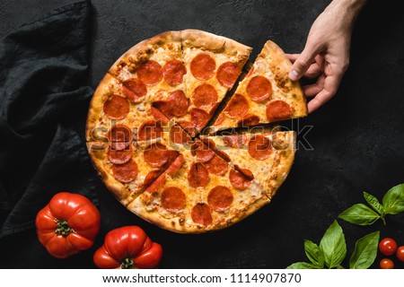 Hand picking slice of pepperoni pizza. Pizza party. Hot italian pepperoni pizza on black concrete background, top view