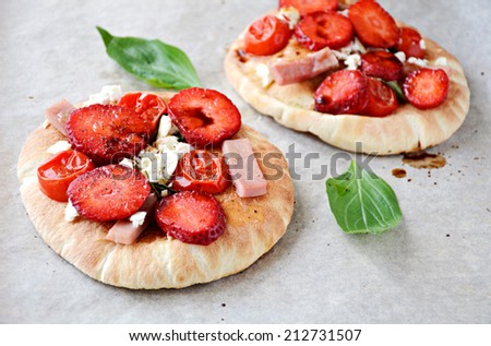 Small pizza on pita bread with tomatoes, ham, white cheese and strawberries topped with balsamic sauce on gray background. Fresh basil leaves