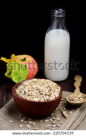 Rolled oats in bowl with bottle of milk, fresh apple, wooden spoon and wheat ear on linen. Vertical food still life