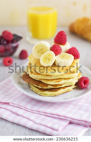 Stack of pancakes with banana and coconut topped with fresh raspberries, coconut flakes and banana slices. Orange juice, flaky croissant, cranberries and dark chocolate on the background
