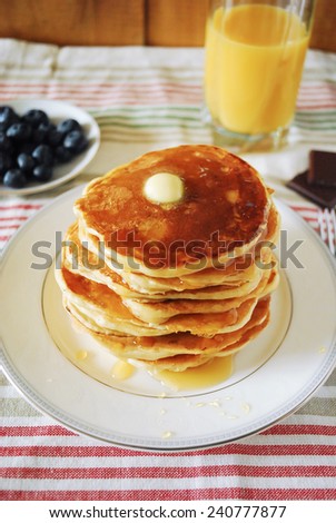 Stack of pancakes with knob of butter and maple syrup on top, served with fresh blueberries and orange juice