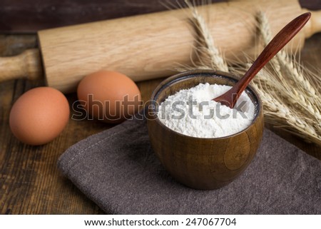 White wheat flour in brown wooden bowl, fresh eggs, wheat ears and rolling pin on wooden table. Cooking at countryside, food ingredients