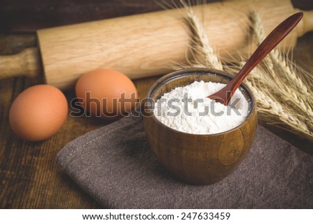 White wheat flour in brown wooden bowl, fresh eggs, wheat ears and rolling pin on wooden table. Cooking at countryside, food ingredients still life