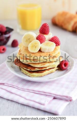 Gourmet breakfast: stack of pancakes with banana and coconut topped with fresh raspberries, coconut and banana. Orange juice, flaky croissant, fresh cranberries and dark chocolate on background