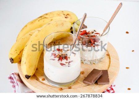 Two glasses with banana yogurt topped with granola, milk chocolate and goji berries on round wooden plank with bananas and milk chocolate bars on red napkin