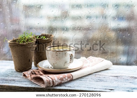 Cup with warm drink on wooden table in front of window with rain drops, rainy weather. Moody still life. Warm tones, horizontal image