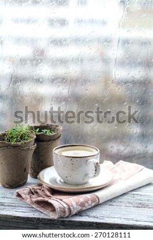 Cup with warm drink on wooden table in front of window with rain drops, rainy weather. Moody still life