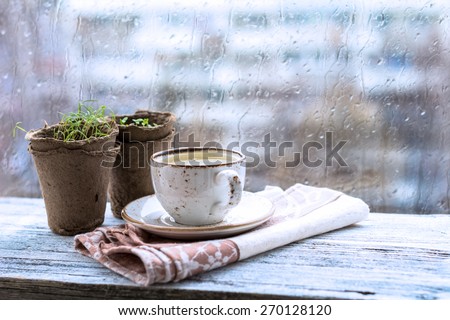 Cup with warm drink on wooden table in front of window with rain drops, rainy weather. Moody still life. Cold tones, horizontal image
