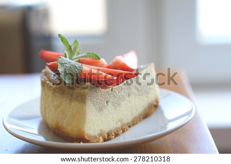 Piece of homemade cheesecake (with matcha green tea layer) decorated with fresh strawberries and mint on edge of kitchen table near window